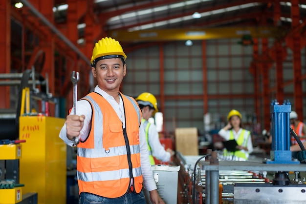 Ingeniero masculino asiático usa chaleco de seguridad con casco amarillo de pie sosteniendo una herramienta de llave en fábrica