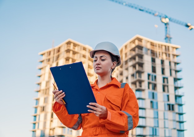 Ingeniero de mujer de negocios hermosa vistiendo un mono naranja está de pie con los documentos del plan. Proyecto de construcción con grúa está en segundo plano.