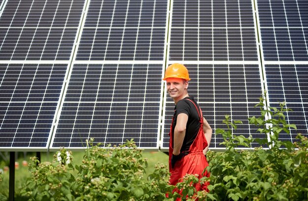 Ingeniero de pie en casco de construcción amarillo frente a la estación de energía solar fotovoltaica en un día soleado con vegetación