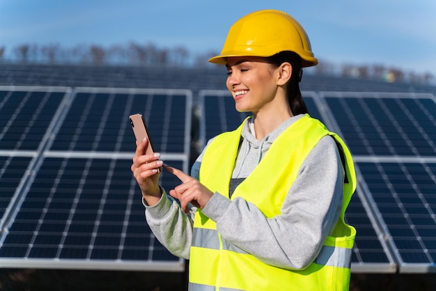 Ingeniero de una planta de energía solar blogueando en la página de la empresa en la red social Mujer joven con chaleco amarillo sosteniendo un teléfono inteligente en las manos publicando contenido nuevo sobre el fondo de los paneles solares