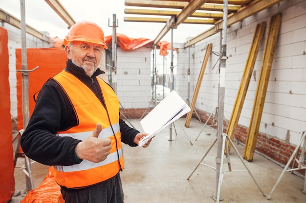 Ingeniero senior o trabajador de la construcción con casco que aprueba la construcción de una nueva casa moderna con planos Retrato de un arquitecto o contratista masculino que tiene planes en el sitio de construcción