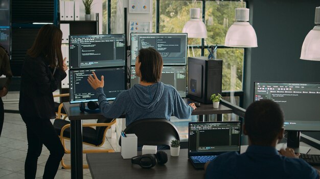 El ingeniero de sistemas verifica el código en varios monitores y trabaja con un colega desarrollador de aplicaciones en la oficina de la agencia de TI. Codificador analizando algoritmos en pantallas, desarrollando una nueva interfaz de usuario.
