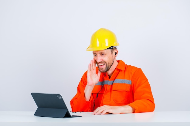 El ingeniero sonriente posa sosteniendo la mano cerca de la boca y sentado frente a su tableta en el fondo blanco