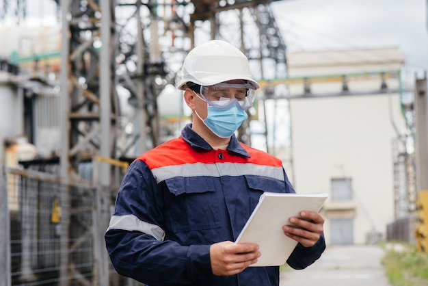 Un ingeniero de subestación eléctrica inspecciona equipos modernos de alto voltaje con una máscara en el momento de la pondemia. Energía. Industria.