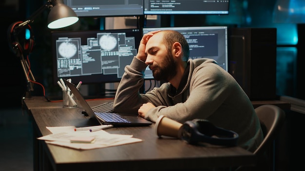 Ingeniero de TI de aplicaciones durmiendo en el escritorio y agotado, tratando de trabajar con inteligencia artificial por la noche. Hombre cansado y soñoliento bostezando y quedándose dormido en la oficina, usando múltiples monitores.