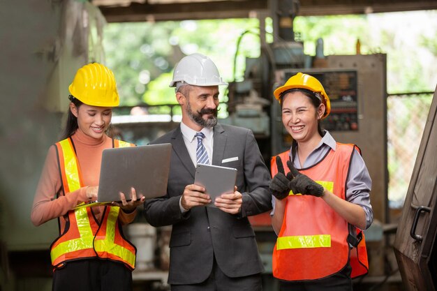 Ingeniero y trabajadores industriales en cascos discuten un nuevo proyecto mientras usan una computadora portátil. Hacen gestos de mostrar Trabajan en una fábrica de fabricación de industria pesada.