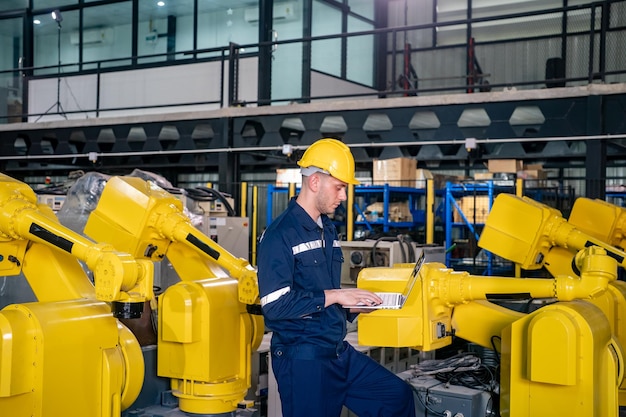 Ingeniero trabajando en una computadora portátil en una instalación industrial con brazos robóticos durante las horas de funcionamiento