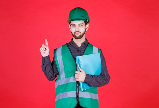 Ingeniero en uniforme verde y casco sosteniendo una carpeta azul y apareciendo.