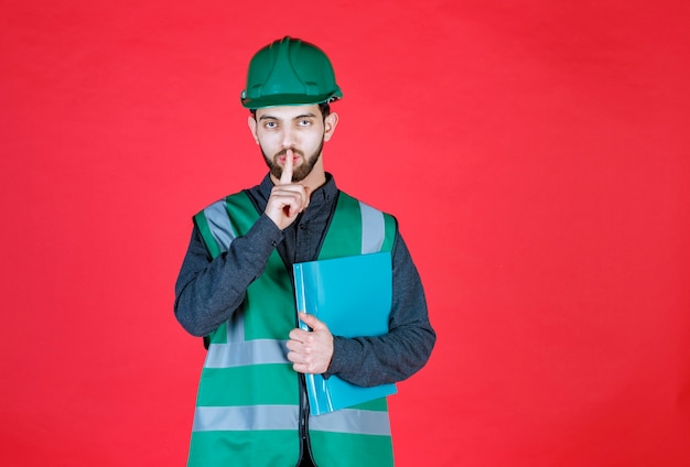 Ingeniero en uniforme verde y casco sosteniendo una carpeta azul y pidiendo silencio.