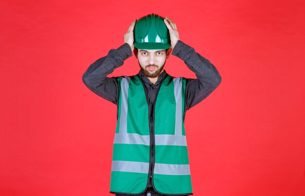 Foto ingeniero en uniforme verde y casco sosteniendo su cabeza porque está cansado.