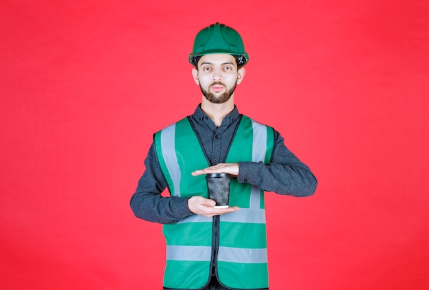 Ingeniero en uniforme verde y casco sosteniendo una taza de café desechable negra.