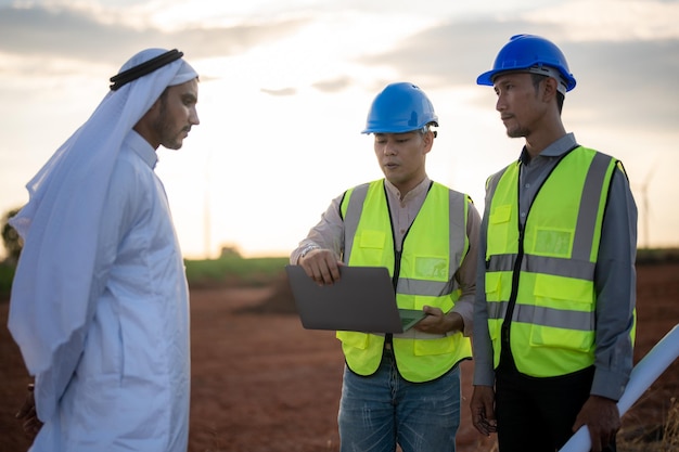 Ingenieros asiáticos y empresarios árabes revisando e inspeccionando la construcción con la operación de personas del cielo del atardecer Turbina eólica para electricidad de energía limpia y medio ambiente Industrial de sostenible