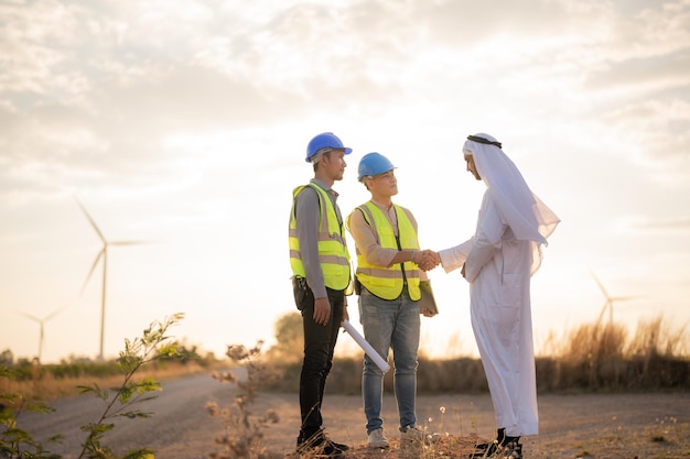 Ingenieros asiáticos y empresarios árabes revisando e inspeccionando la construcción con la operación de personas del cielo del atardecer Turbina eólica para electricidad de energía limpia y medio ambiente Industrial de sostenible