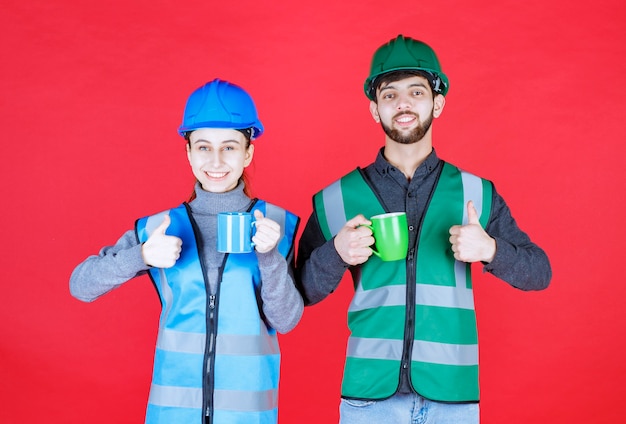 Ingenieros masculinos y femeninos con casco sosteniendo tazas azules y verdes y mostrando el signo de satisfacción.