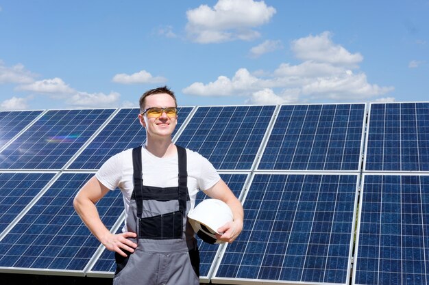 Ingenieur für Sonnenkollektoren in gelben Schutzgläsern und grauen Overalls, die in der Nähe des Feldes für Sonnenkollektoren stehen und ein weißes Fass halten