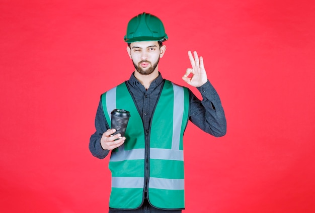 Ingenieur in grüner Uniform und Helm, der eine schwarze Einwegtasse Kaffee hält und den Geschmack genießt.