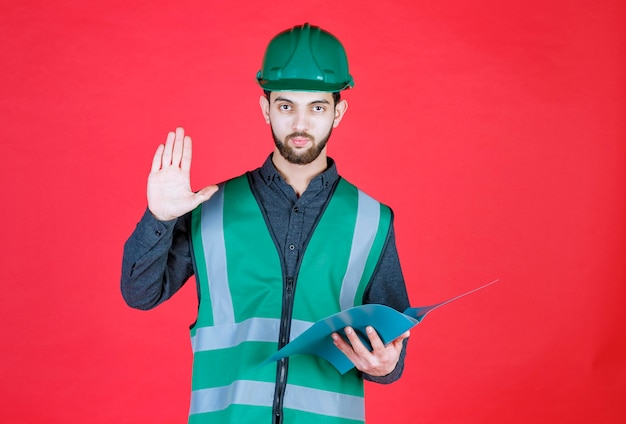 Ingenieur in grüner Uniform und Helm, der einen blauen Ordner hält, liest und korrigiert.