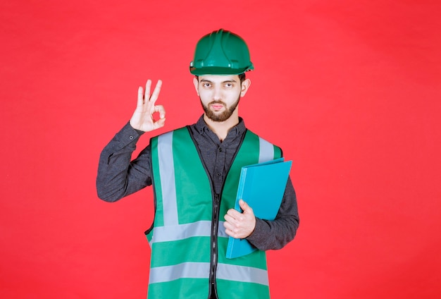 Ingenieur in grüner Uniform und Helm, der einen blauen Ordner hält und ein positives Handzeichen zeigt.