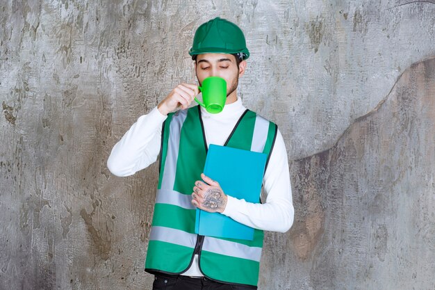 Ingenieur Mann in der gelben Uniform und im Helm, die eine grüne Kaffeetasse und eine grüne Mappe halten.