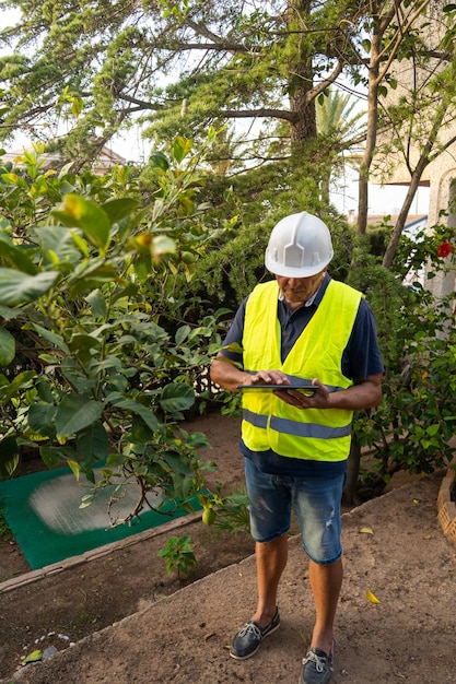Ingenieur mit einem elektronischen Gerät inspiziert einen Hausgarten auf Schädlinge Kontrollen auf dem Tablett trägt eine Weste und einen Helm