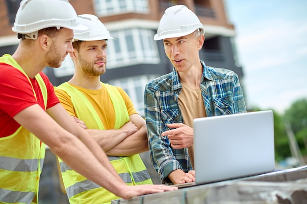 Ingenieur und zwei Arbeiter diskutieren im Freien in der Nähe eines Laptops