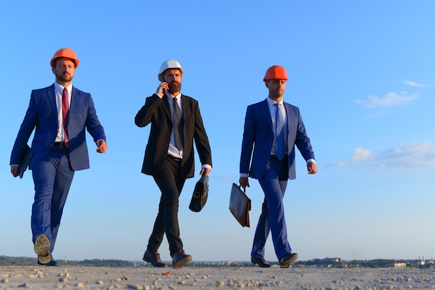 Ingenieure halten schwarze und braune Koffer. Architekten mit strengen Gesichtern in Anzügen, weißen und orangefarbenen Helmen. Bauherren gehen selbstbewusst und sprechen am Telefon auf blauem Himmelshintergrund. Geschäfts- und Gebäudekonzept