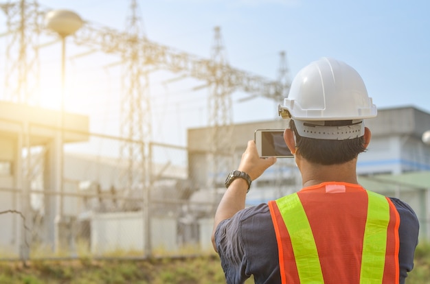 Ingenieure verwenden Mobiltelefone, um Hochspannungsmasten vor dem Hintergrund eines Umspannwerks zu fotografieren.