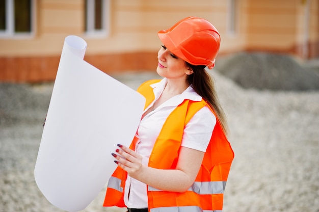 Ingenieurerbauerfrau in der einheitlichen Weste und im orange Schutzhelm halten Geschäftspapier gegen Neubau