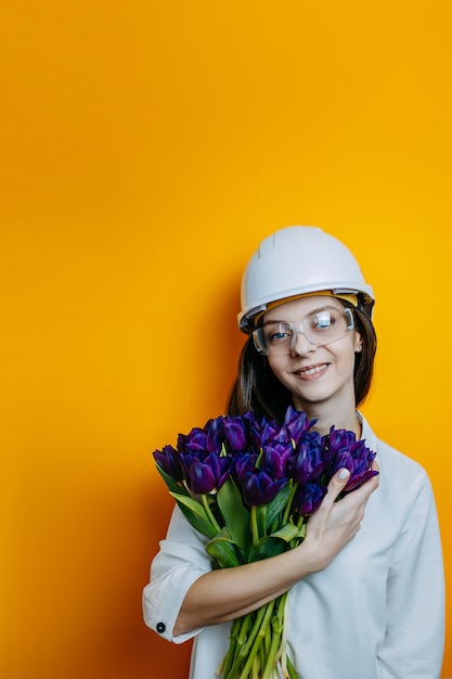 Ingenieurin in weißem Helm und Brille. Sicherheitsplakat. Frau im weißen Hemd hält großen Bündel lila Tulpen.