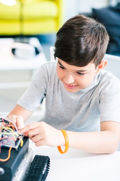 Ingenioso colegial encantado sentado a la mesa y construyendo un dispositivo robótico