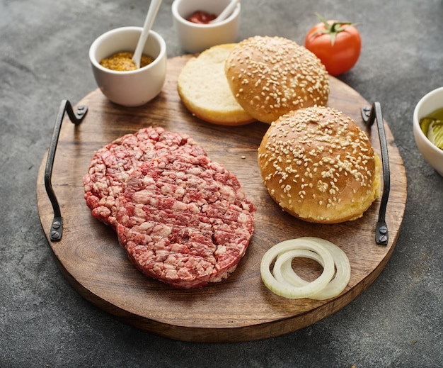 Los ingredientes crudos para la hamburguesa casera en tablero de madera Empanadas de hamburguesa Carne picada cruda chuleta molida de res y cerdo con panecillo Fondo gris