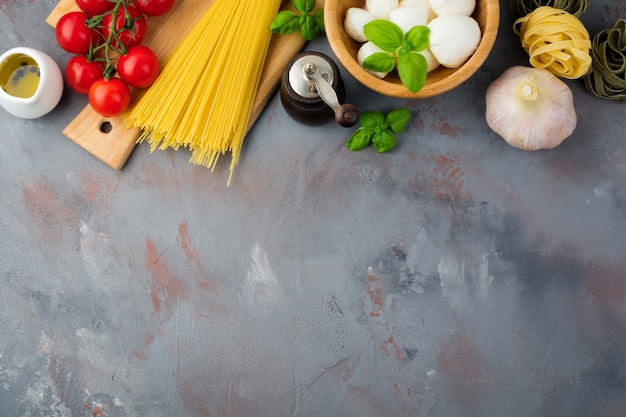 Ingredientes crudos para la preparación de pasta italiana, espaguetis, tagliatelle, fusilli, ajo, albahaca, mozzarella, lechuga, pimiento, tomates cherry y aceite de oliva sobre una mesa de hormigón gris. Vista superior