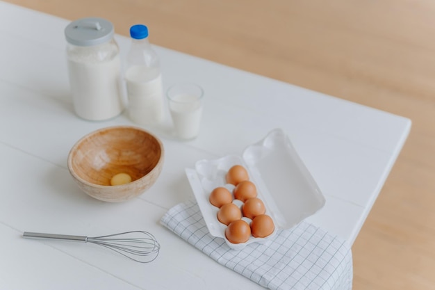 Ingredientes de cozimento na mesa branca. Ovos, leite e farinha em recipientes. Batedor para bater e fazer a massa. Processo de assar. Hora de cozinhar