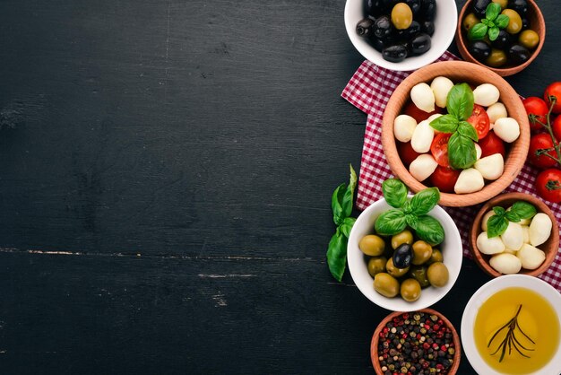 Ingredientes para ensalada caprese italiana Queso mozzarella Tomates cherry Hojas de albahaca Aceitunas Aceite Pimienta Sobre un fondo de madera negra Espacio libre para texto