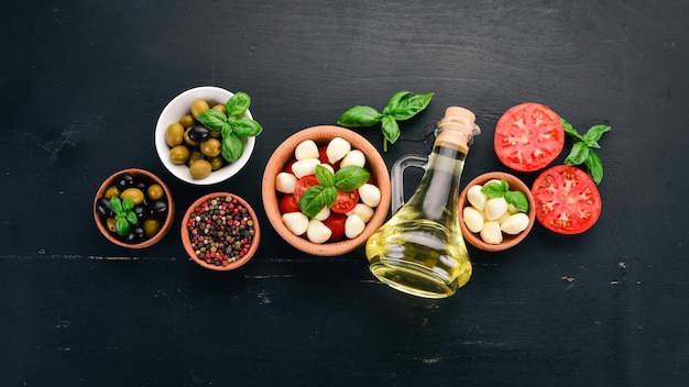 Ingredientes para ensalada caprese italiana Queso mozzarella Tomates cherry Hojas de albahaca Aceitunas Aceite Pimienta Sobre un fondo de madera negra Espacio libre para texto