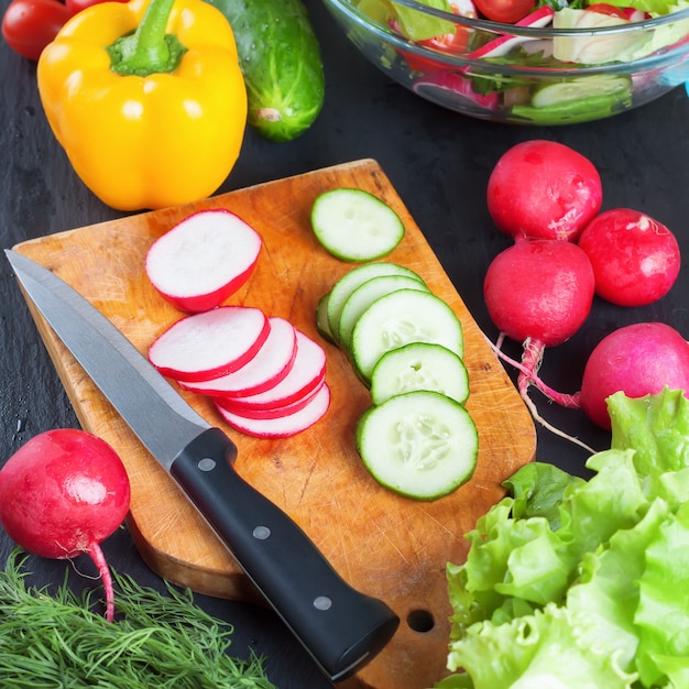 Ingredientes para la ensalada en rodajas sobre tabla de cortar con cuchillo. verduras frescas sobre fondo de pizarra negra. vista superior.