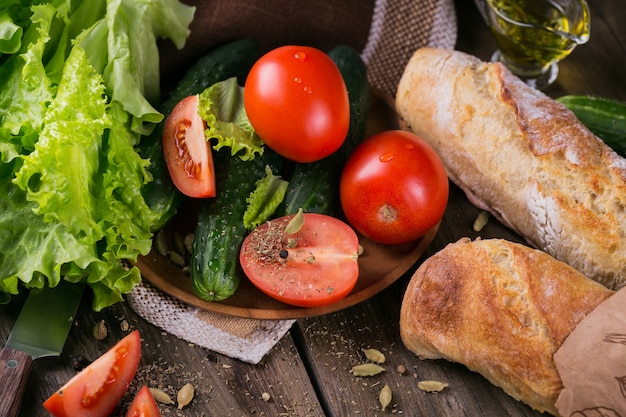 Ingredientes frescos para cozinhar salada: tomate, pepino, alface, azeite e especiarias sobre fundo de mesa de madeira rústica. Servido com baguete fresca. Conceito de alimentação saudável