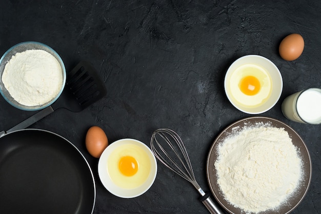 Ingredientes para assar panquecas. frigideira, ovos, farinha, batedor de ovos, em um preto