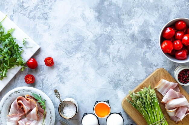 Ingredientes para cozinhar em um fundo cinza de concreto. Um monte de aspargos verdes frescos, ovos, bacon, tomate, rúcula. Vista do topo. Copie o espaço