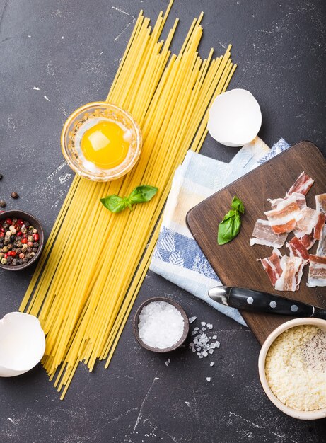 Ingredientes para cozinhar macarrão italiano tradicional espaguete carbonara, gema crua, queijo parmesão, bacon na placa de madeira sobre fundo preto de pedra rústica