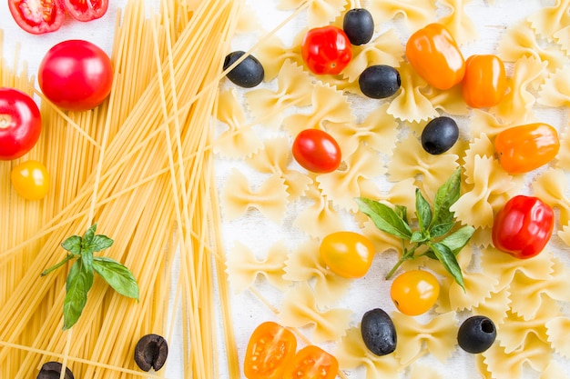 Ingredientes de la pasta, pasta cruda, tomates cherry, aceitunas y hojas de albahaca sobre el fondo blanco.