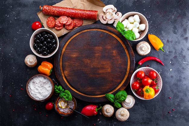 Ingredientes de pizza en el fondo oscuro y tabla de cortar redonda. Pepperoni, mozzarella, tomates, aceitunas, champiñones y harina son productos diferentes para hacer pizza y pasta.