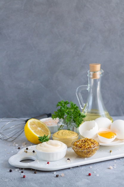 Ingredientes para la salsa de mayonesa casera: mostaza, huevos, aceite de oliva, limón, especias y hierbas sobre fondo de piedra.