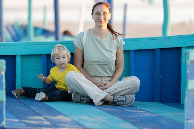 Ingwerfrau mit Brille und ihr kleiner blonder Sohn sitzen auf den blauen Brettern am Strand