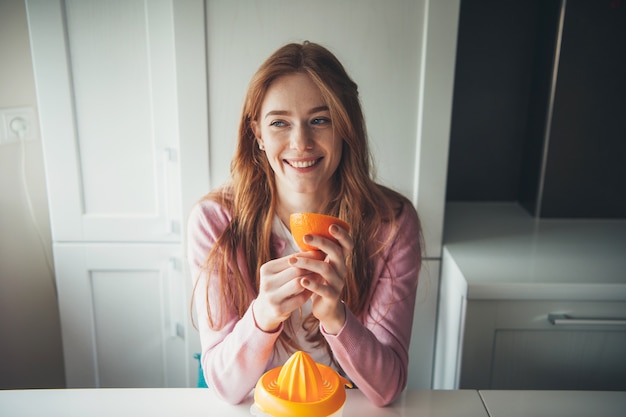 Ingwerfrau mit Sommersprossen lächelt, während sie eine geschnittene Orange hält, während sie Fruchtsaft drückt