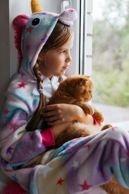 Ingwerkätzchen und ein kleines Mädchen sitzen auf der Fensterbank und schauen aus dem Fenster.