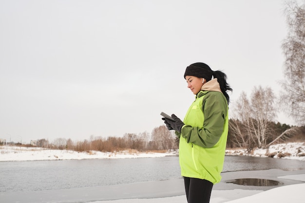 Inhalt attraktiver weiblicher Jogger in den Ohrknospen, die am Fluss stehen und Fitness-App am Telefon überprüfen