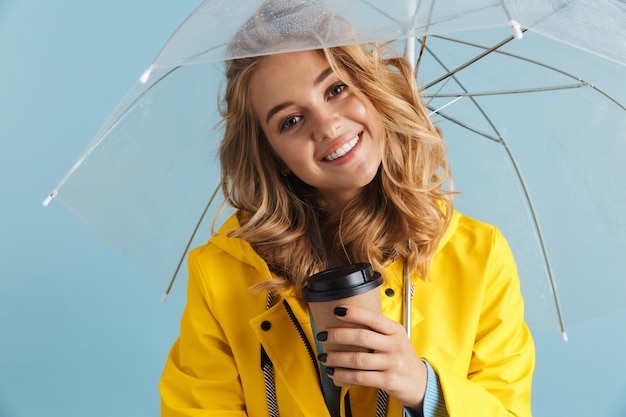 Inhalt Frau 20s tragen gelben Regenmantel stehend unter transparentem Regenschirm mit Kaffee zum Mitnehmen