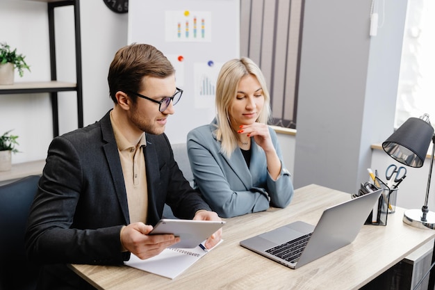 Inicie un equipo de negocios para reunirse en el interior de una oficina moderna trabajando en una computadora portátil y una tableta Una mujer joven y un hombre están sentados a la mesa y trabajando en un proyecto empresarial