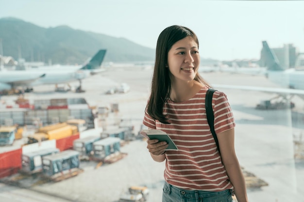 Início da viagem de férias dos jovens. Linda mulher asiática em pé perto da janela enquanto espera o embarque em aeronaves no saguão do aeroporto. senhora sorrindo segurando passaporte e cartão de embarque animado.